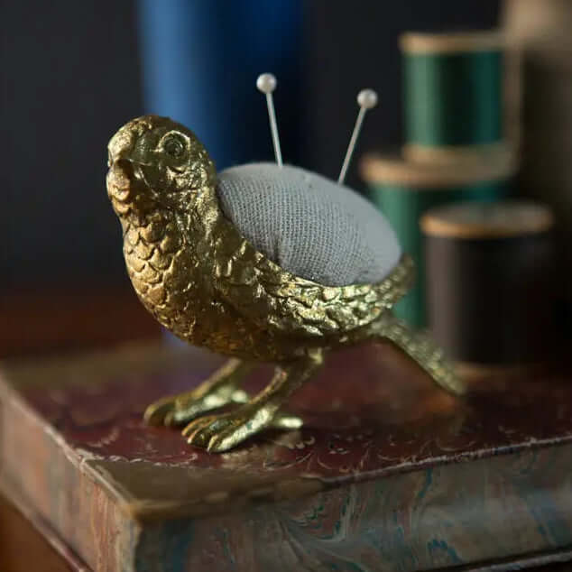Gold-tone pewter bird pincushion beside thread spools and books