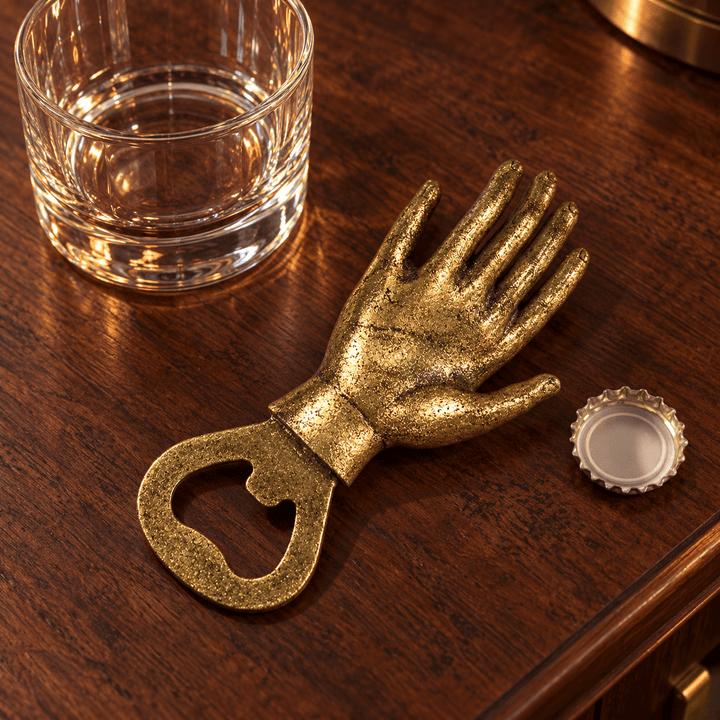 Gold-leaf hand bottle opener on walnut beside a tumbler and bottle cap