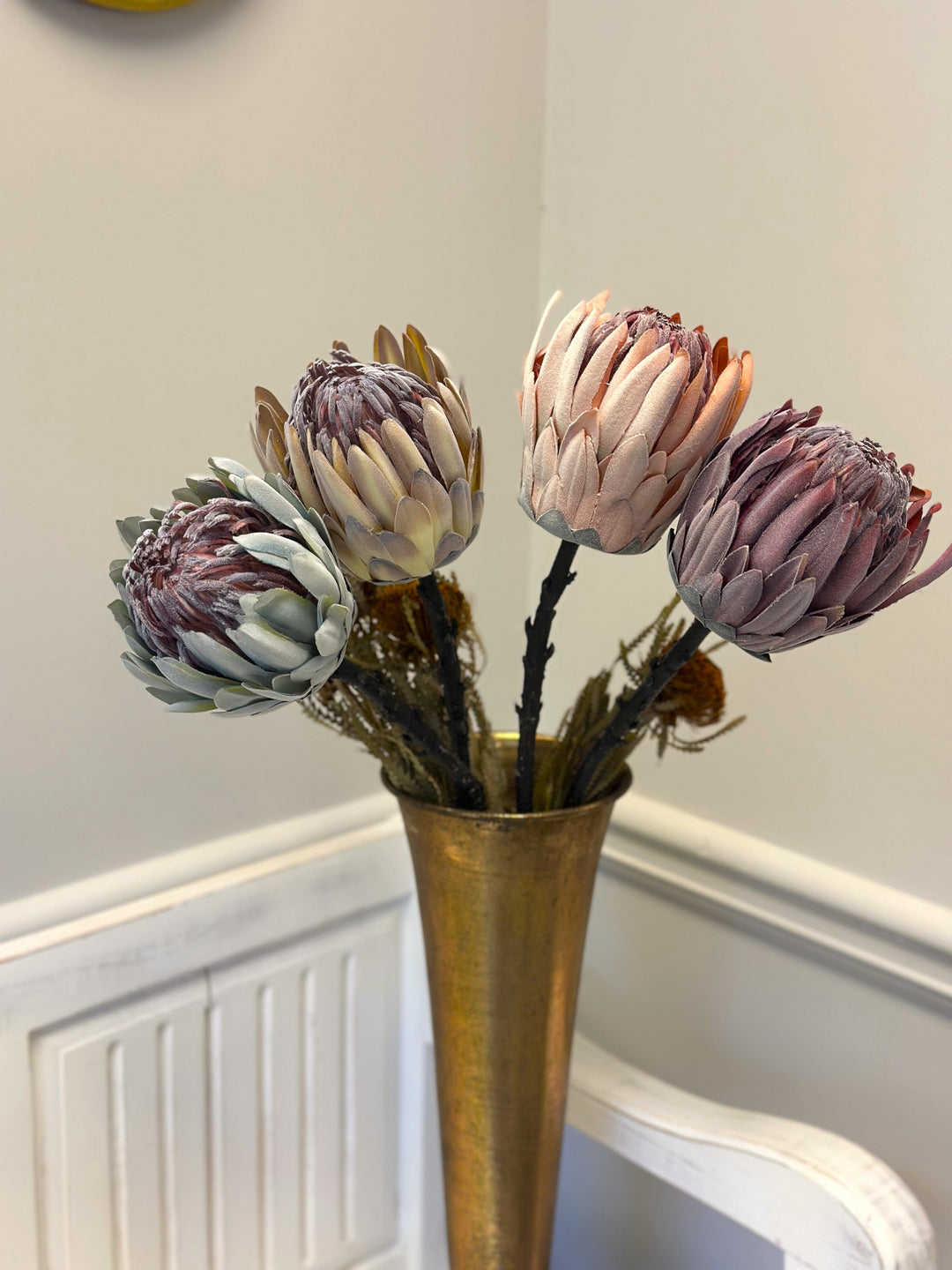 Four king protea faux flower stems displayed together in a tall gold vase