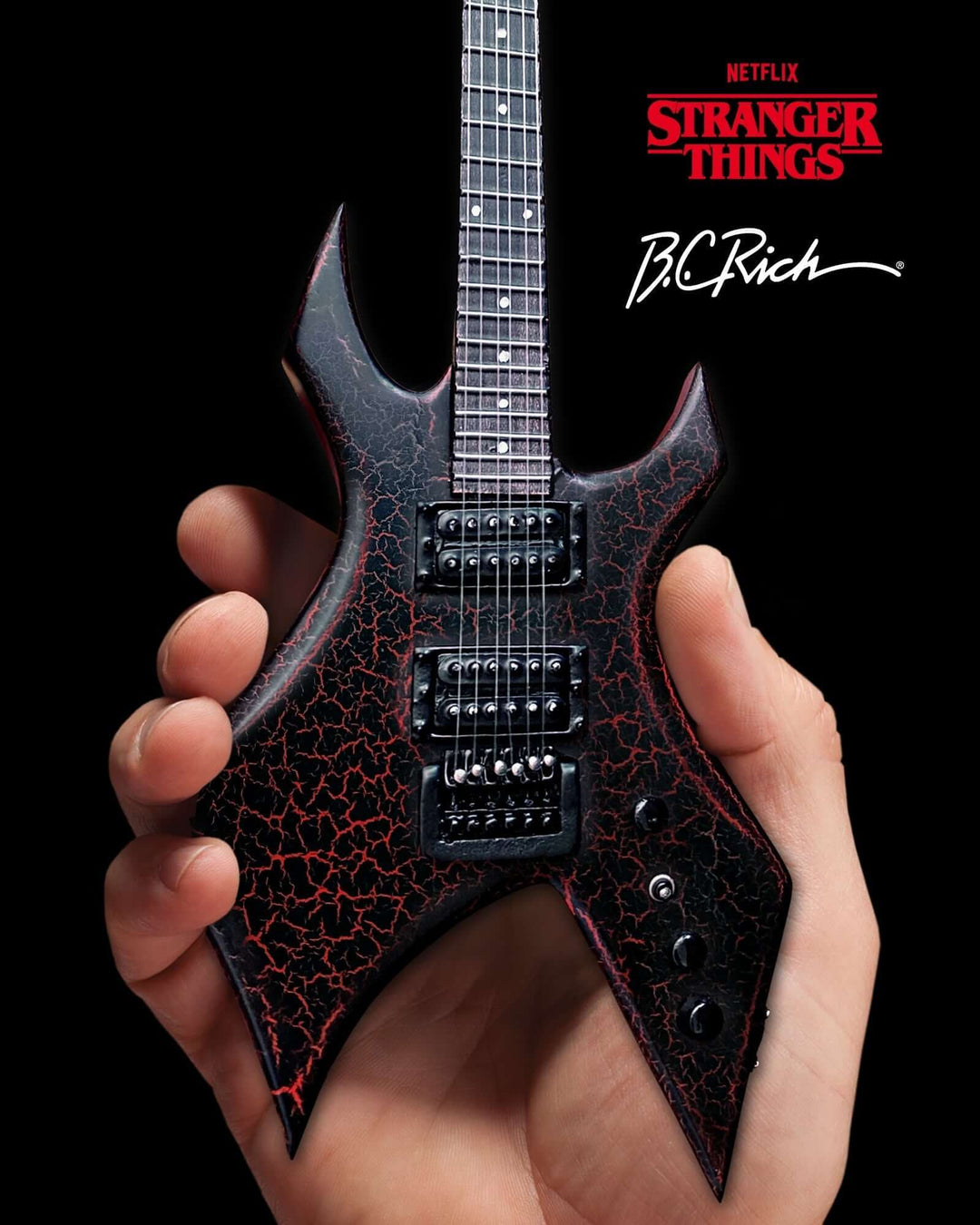 Hand holding Stranger Things Eddie’s B.C. Rich Warlock Mini Guitar Replica against a black background, featuring cracked red design.