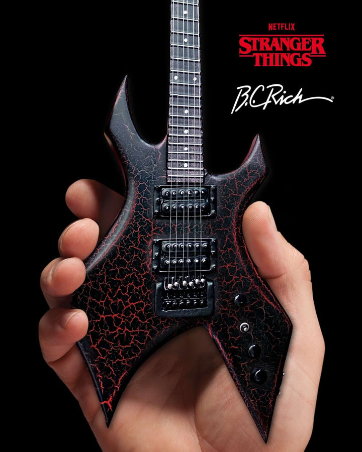 Hand holding Stranger Things Eddie’s B.C. Rich Warlock Mini Guitar Replica against a black background, featuring cracked red design.
