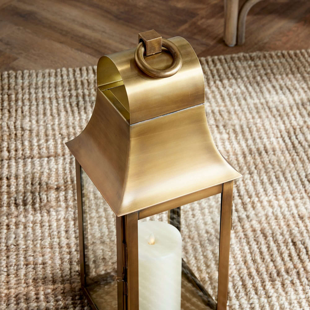 Tall antique brass candle lantern with hanging loop, evoking old-world charm and elegance, featuring a luminous finish and vintage design.