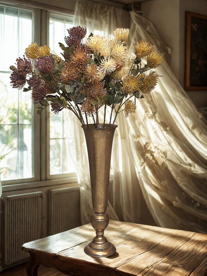 Mixed pincushion protea faux flower stems in a tall antique gold vase by a window
