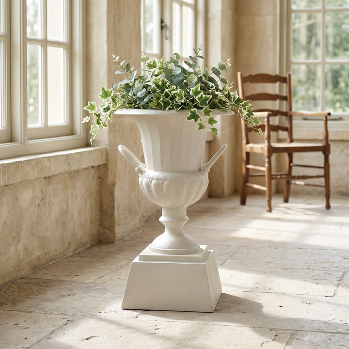 Westbury Iron Urn Planter with Pedestal in Soft White, styled with greenery in a sunlit room beside tall windows.
