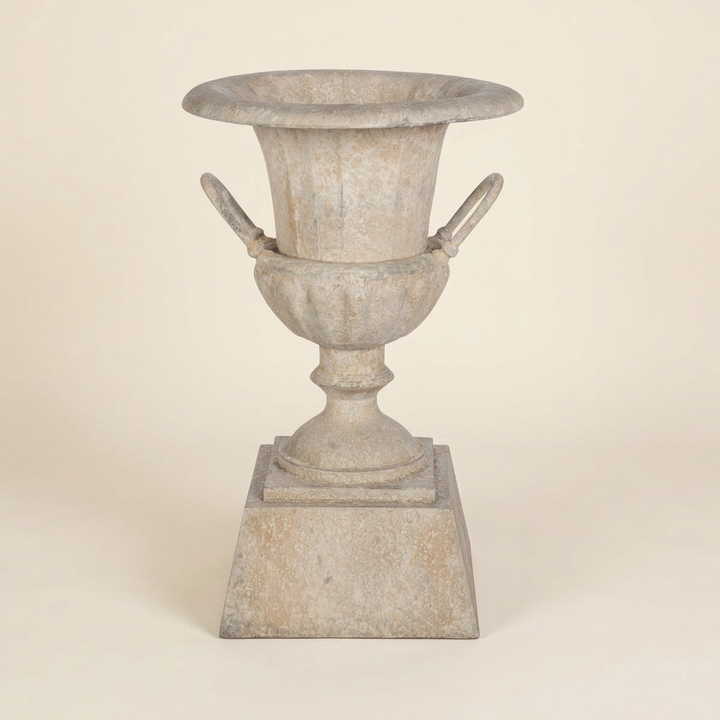Westbury Iron Urn Planter with Pedestal in Weathered Stone, shown empty against an ivory studio background with loop handles and square base visible.