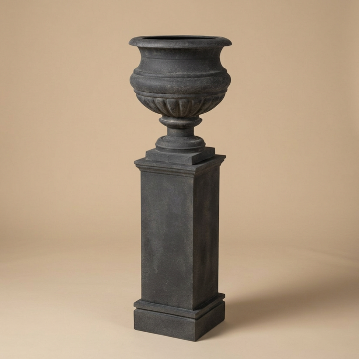 Montclair Iron Urn with Pedestal in Black against a warm beige studio background, showing the flared urn and tall square pedestal.