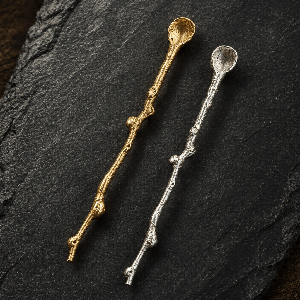 Close-up of gold and silver decorative serving spoons with textured twig-shaped handles on charcoal slate.