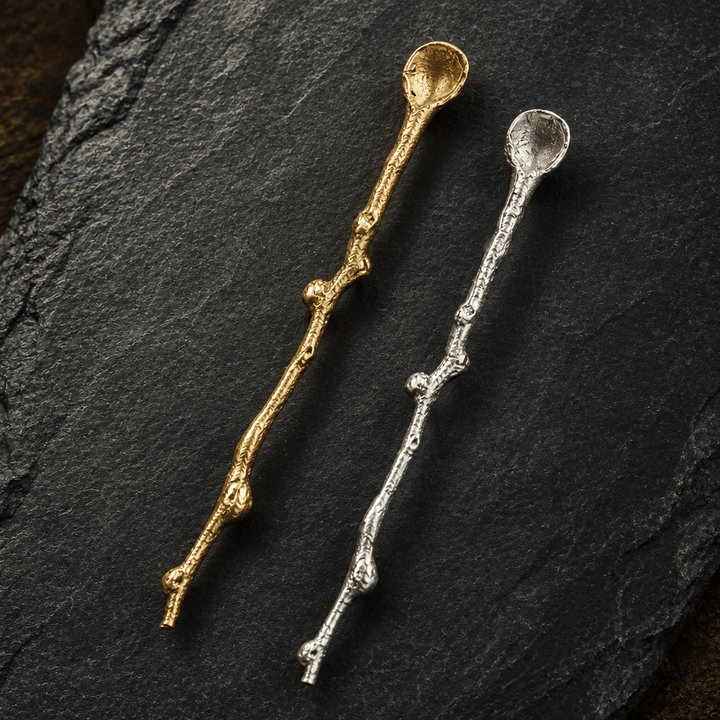 Close-up of gold and silver decorative serving spoons with textured twig-shaped handles on charcoal slate.