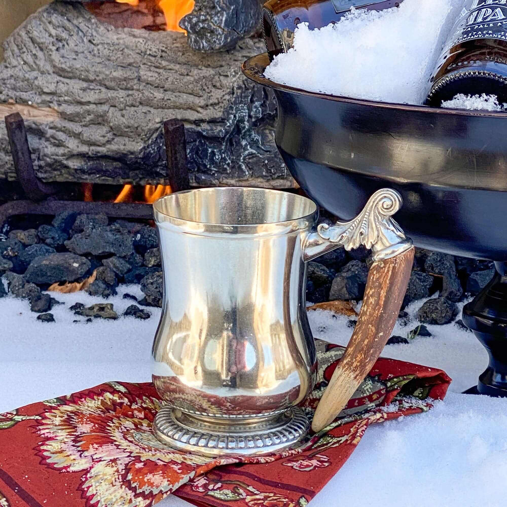 Silver mug with horn handle on a red patterned cloth in front of a fireplace.