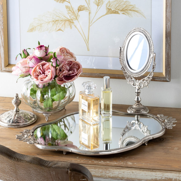Elegant vintage vanity setup with ornate silver tray, perfume bottles, pink roses in glass vase, and antique mirror