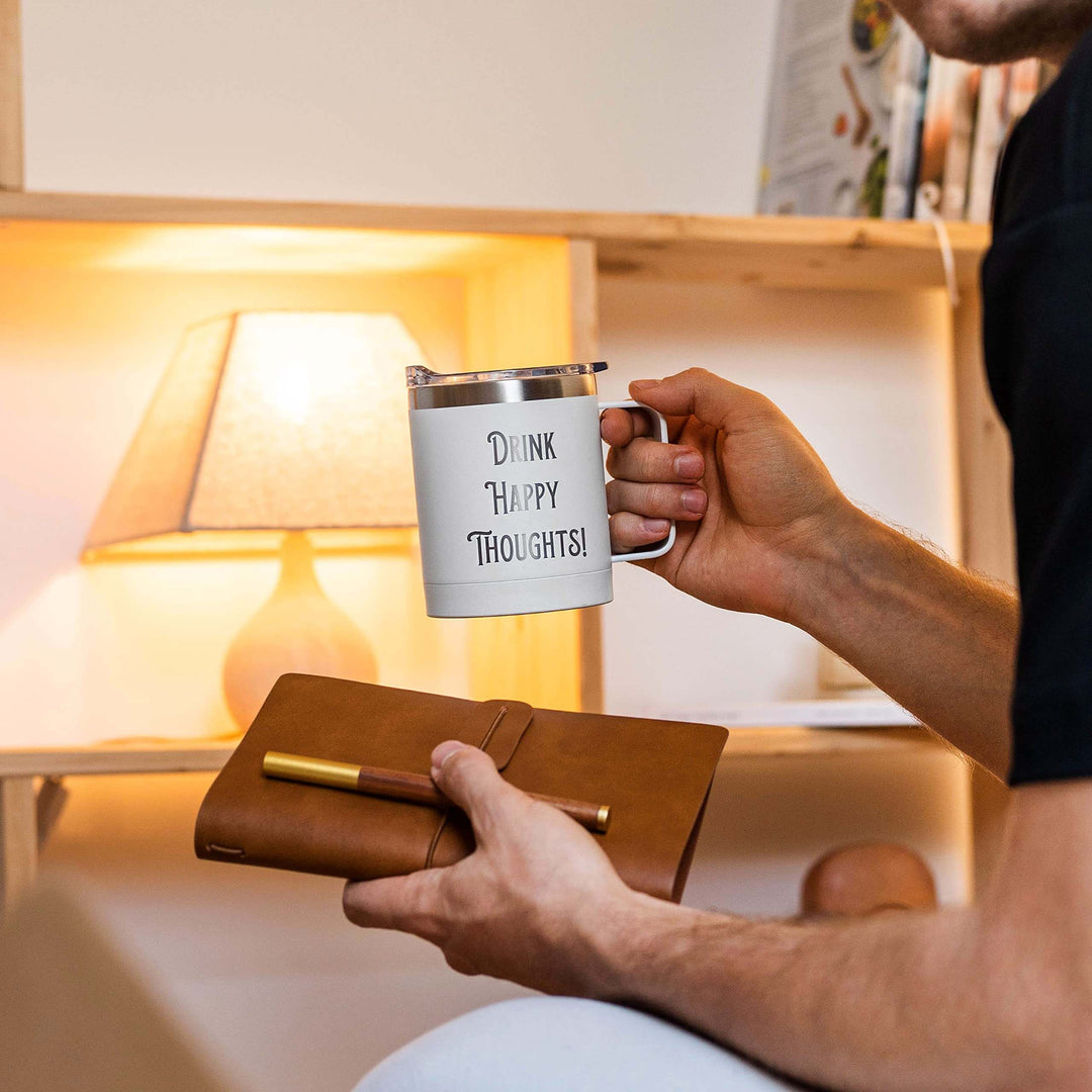 Man holding a mug with 'Drink Happy Thoughts' near a lit lamp and a notepad.