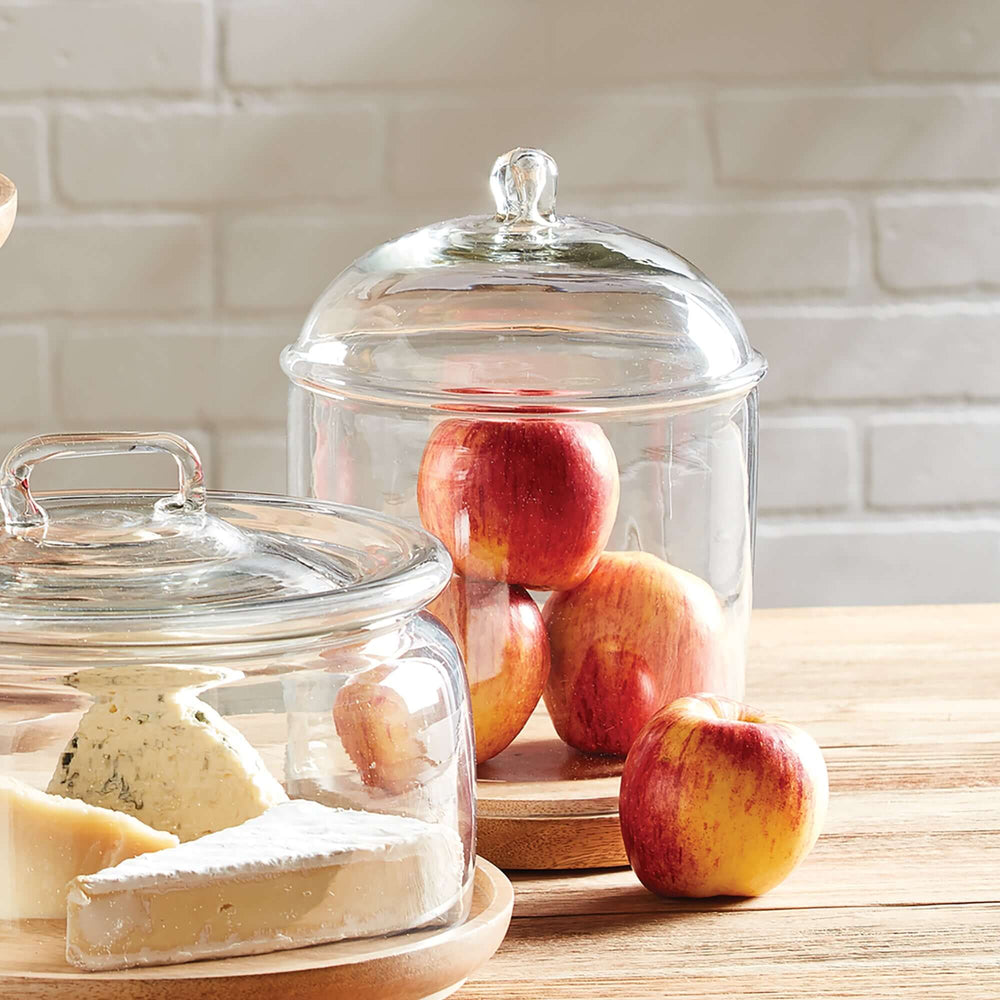 Vintage glass cloche with wood base showcasing apples and cheese on a wooden table.