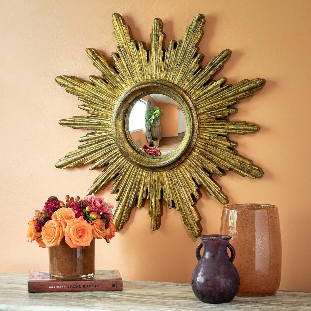 Vintage gold sunburst wall mirror with textured rays and round center, displayed on peach wall with pottery and flowers.