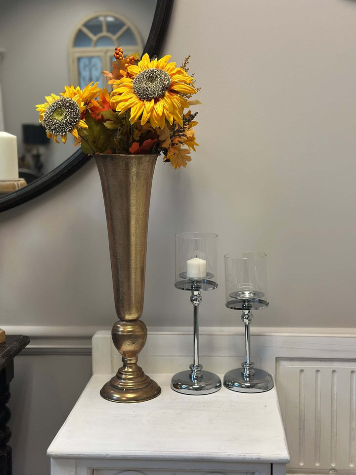 Vintage tall gold trumpet vase with colorful floral arrangement next to glass candle holders on a white table.