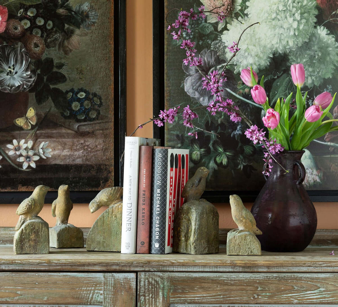 Vintage-inspired bird figurines with aged finish, set of 5, displayed on a rustic shelf beside books and a vase with flowers.