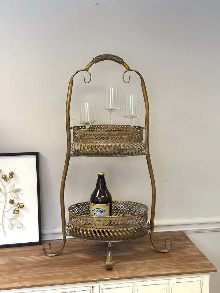 Vintage brass 2-tier metal bar cart with ornate detailing, holding wine glasses and a bottle, on a wooden surface.