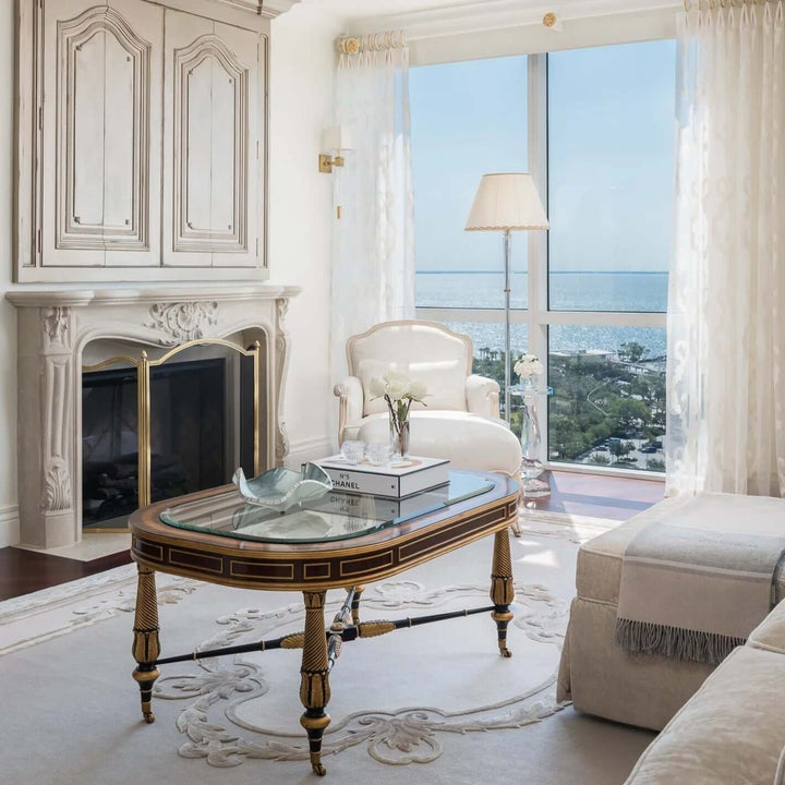 Elegant living room with vintage brass fireplace screen with black mesh, glass coffee table, and ocean view through large windows.