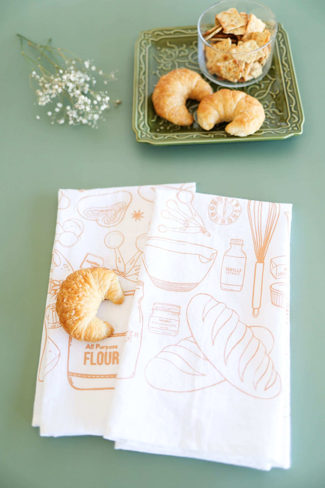 Vintage Bread Baking Tea Towel with hand-drawn illustrations, croissants, and a floral arrangement on a green table.