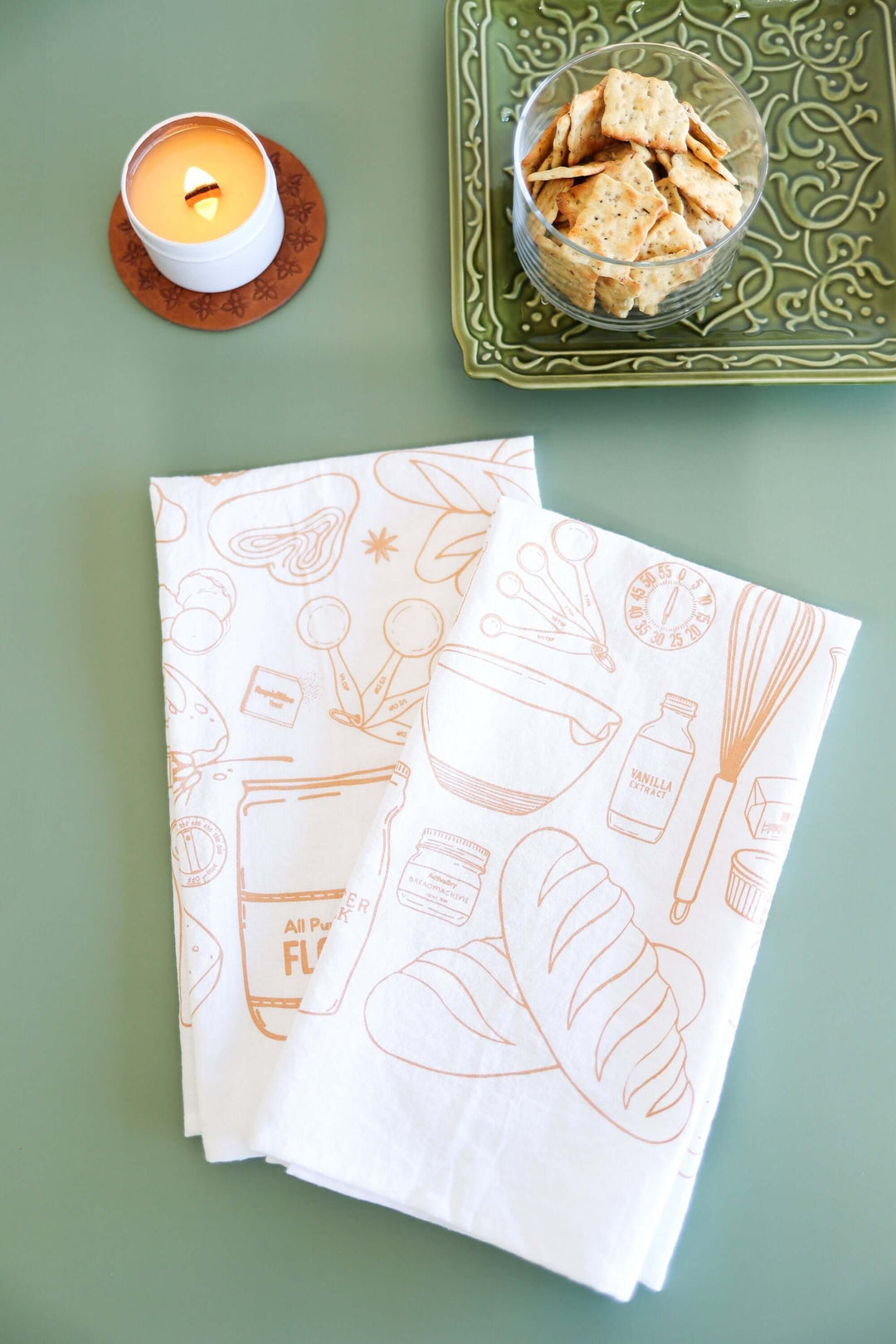 Vintage Bread Baking Tea Towel with hand-drawn illustrations on a kitchen counter, next to a candle and a dish of crackers.