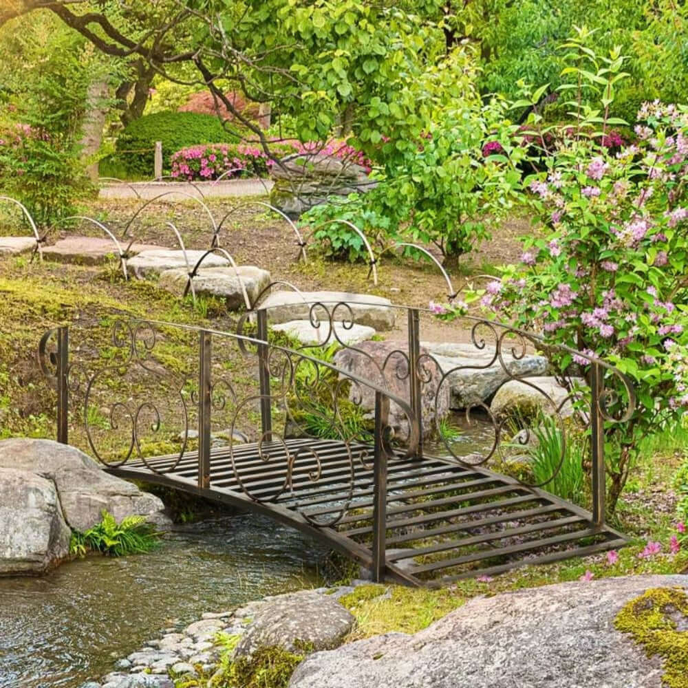 Vintage Bronze Arch Garden Bridge with Scrollwork over a stream in lush garden setting