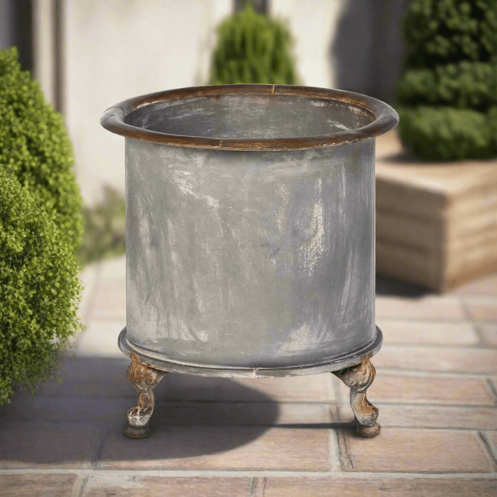 Vintage Creamery Tank Footed Planter made of galvanized iron with rustic patina, styled outdoors with greenery in background.
