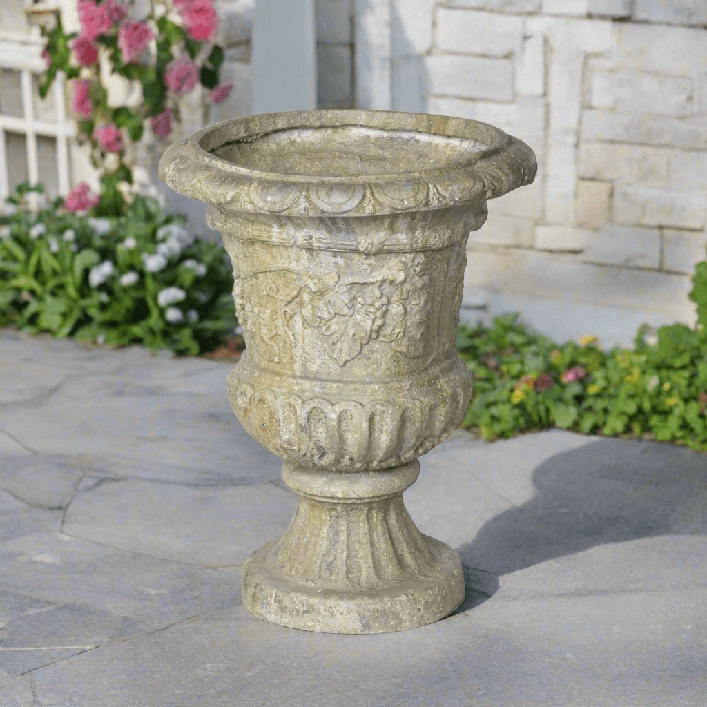 Vintage European Ornate Garden Urn with grapevine carvings, standing on a stone path, surrounded by pink and white flowers.