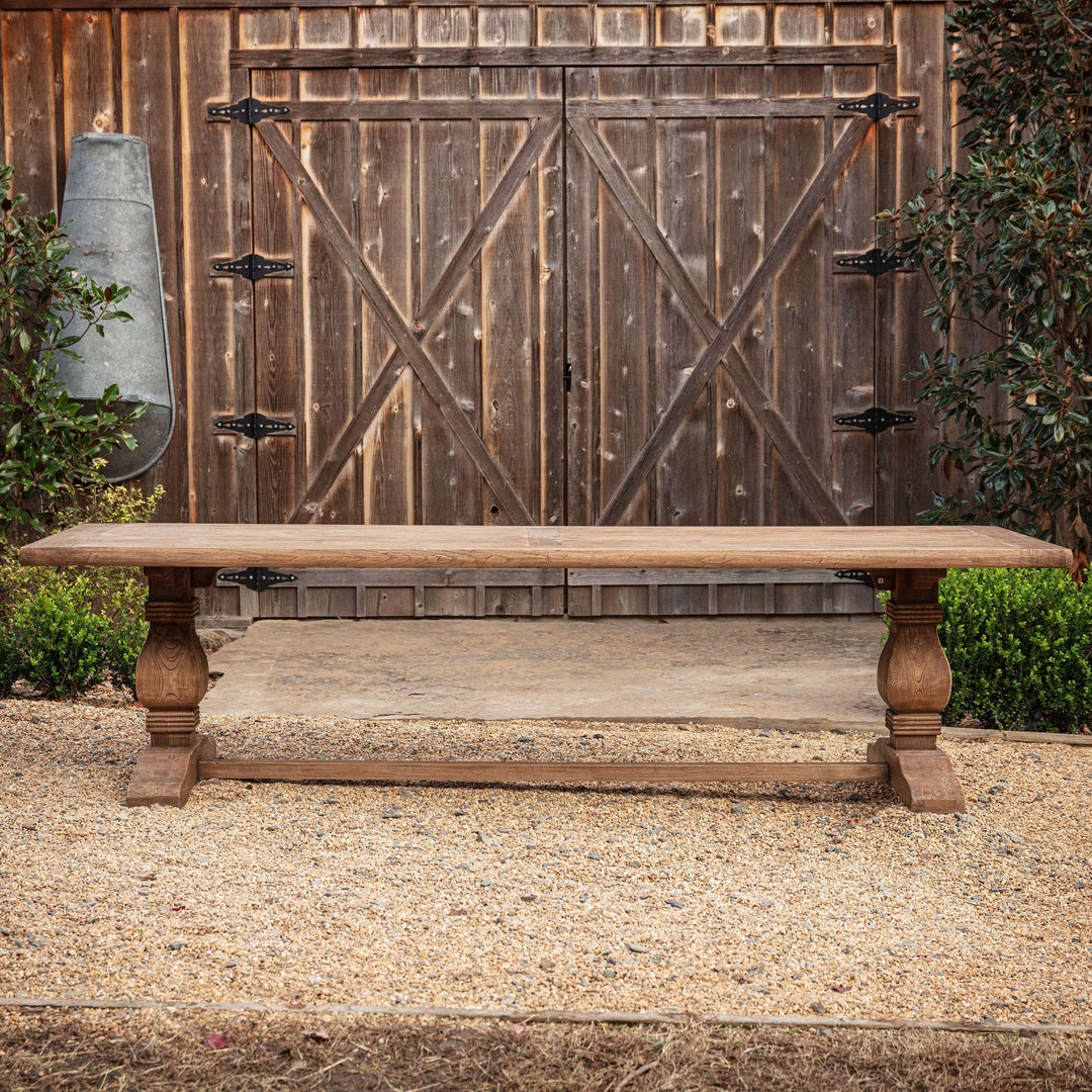 Vintage Farmhouse Harvest Dining Table with rustic wood finish displayed outdoors in a garden setting.