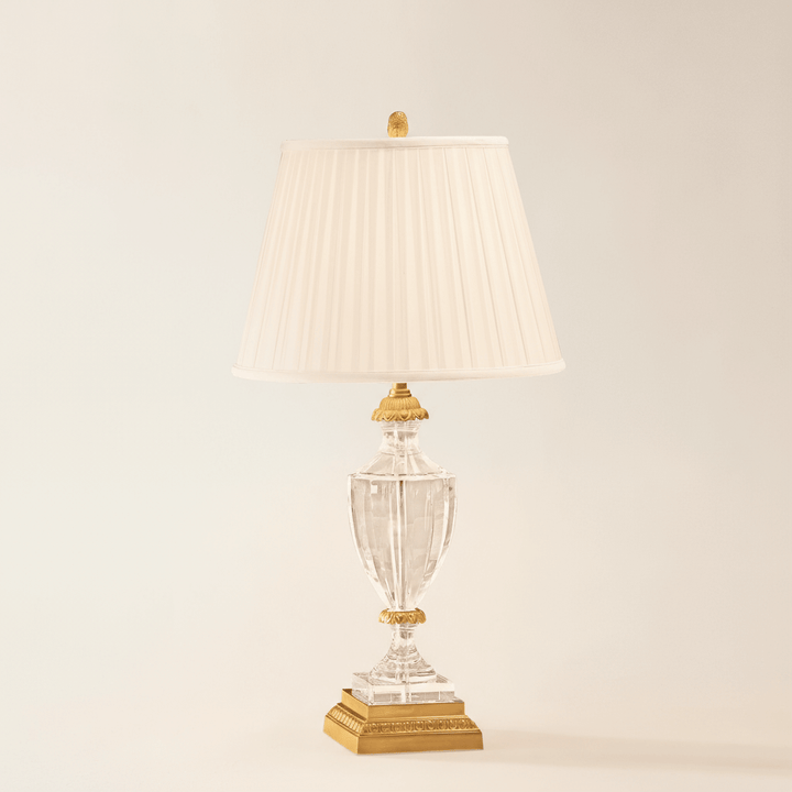 Decorative table lamp with clear glass base and pleated white shade on a beige background
