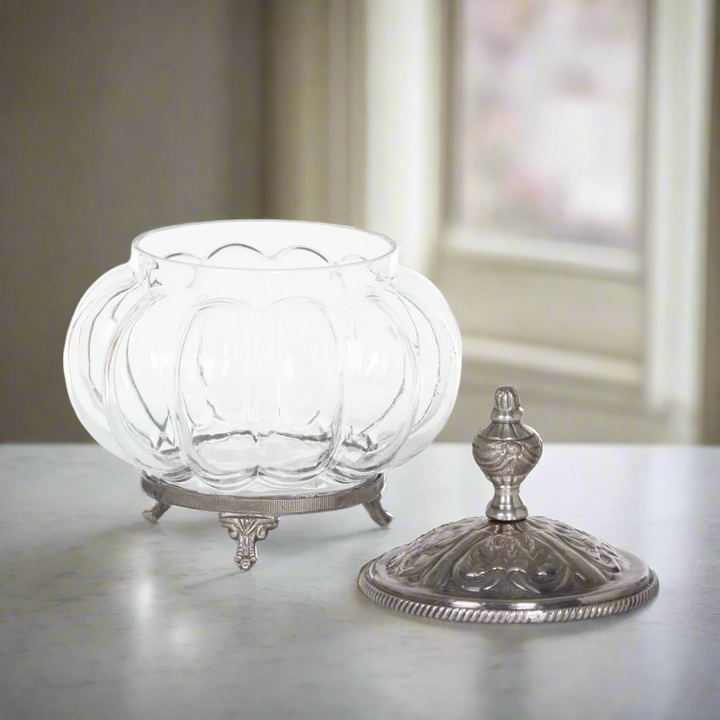 Vintage glass vanity jar with ornate silver lid on display with a pumpkin shape and decorative feet, evokes Victorian elegance.