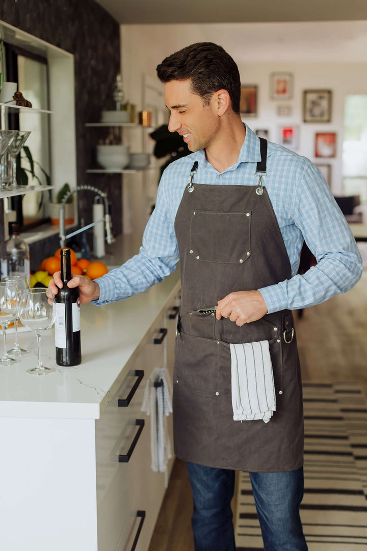 Person wearing Waxed Canvas Bartender Apron and Tools, holding wine bottle in modern kitchen.