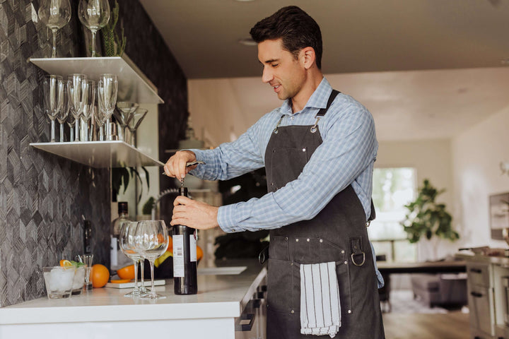 Man using Waxed Canvas Bartender Apron and Tools to open wine bottle in stylish kitchen.