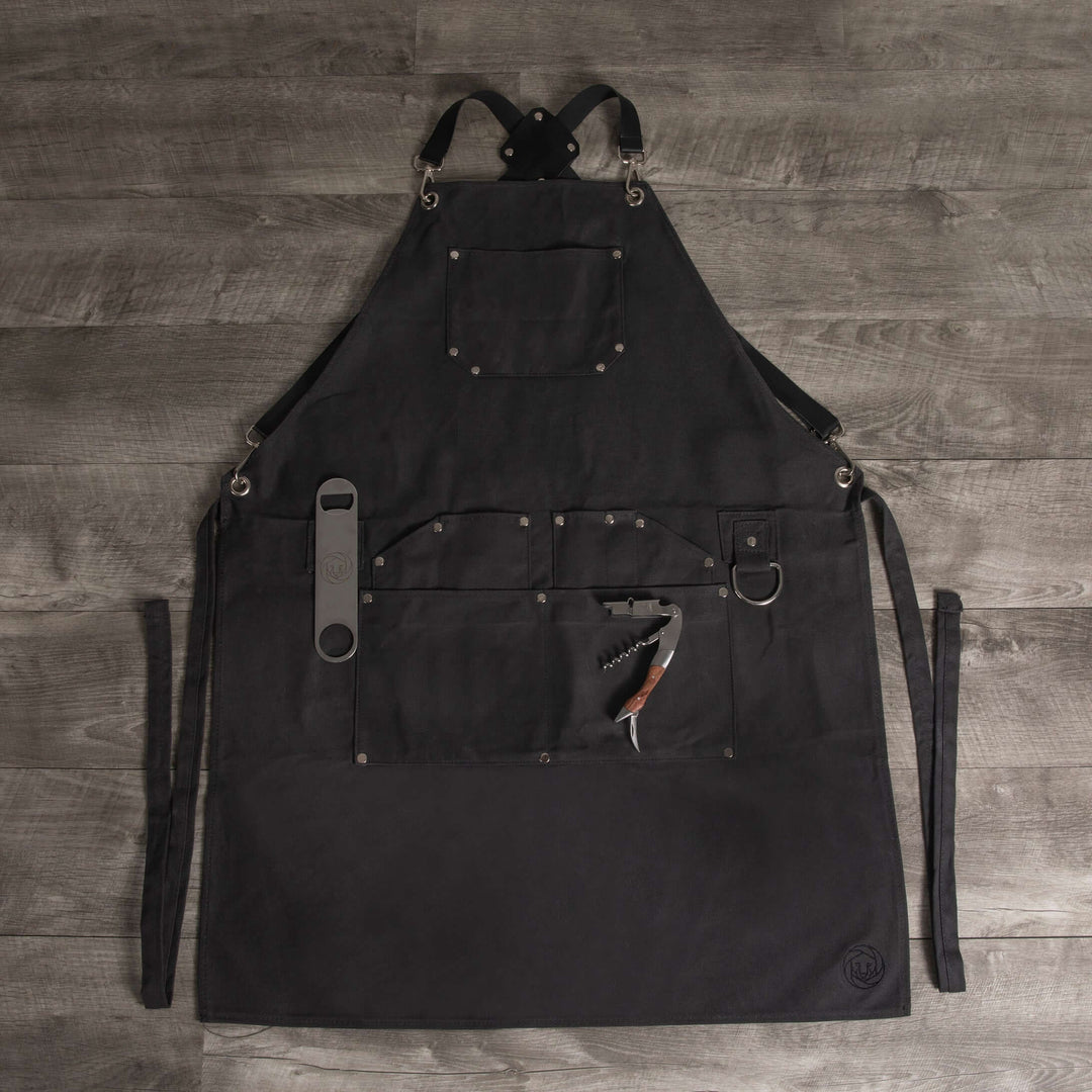 Waxed Canvas Bartender Apron and Tools laid out on wooden floor, featuring pockets and straps for mixology essentials.