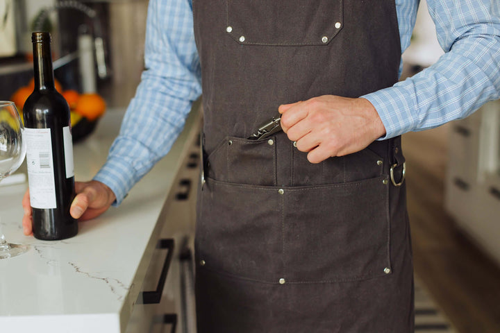 Person wearing Waxed Canvas Bartender Apron and Tools opening wine bottle in kitchen